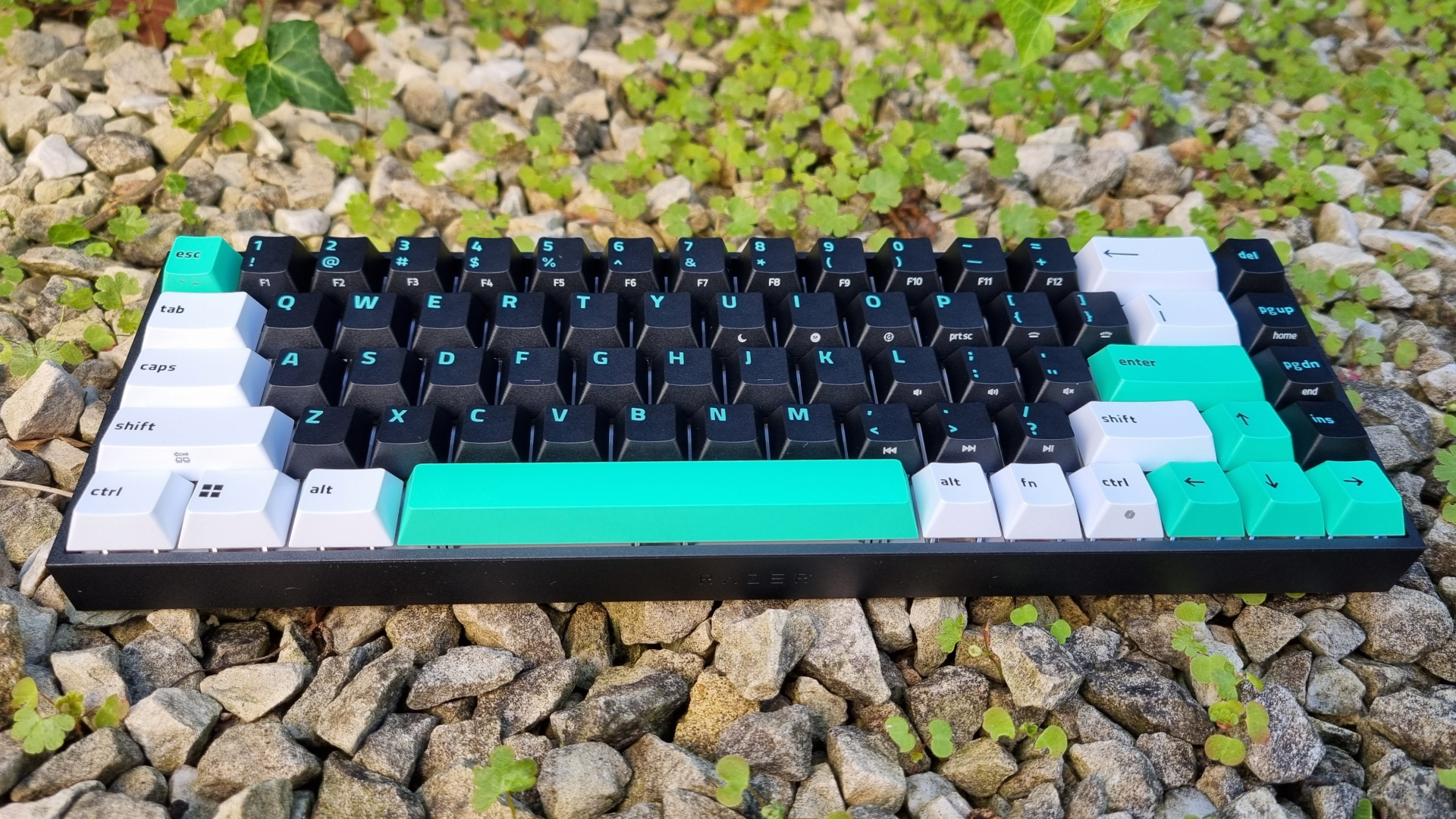 A Razer Reclusa X Mini 65% gaming keyboard on gravel, shot from the user typing position