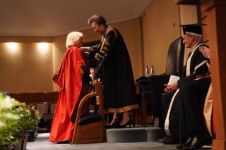 Princess Anne bending forward to give Queen Camilla, who is wearing a red university robe a degree