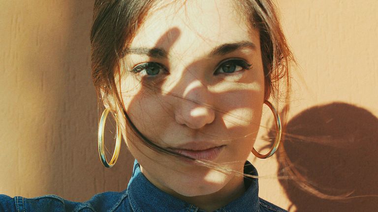 A young woman wearing hoop earings standing against wall holds her hand up to the sun which casts a shadow of her hand on her face