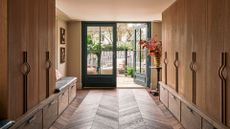 A room with herringbone wood flooring, wooden cabinets and pull-out storage, a storage bench, a vase of stems on a pedestal and green doors leading to an outdoor seating area