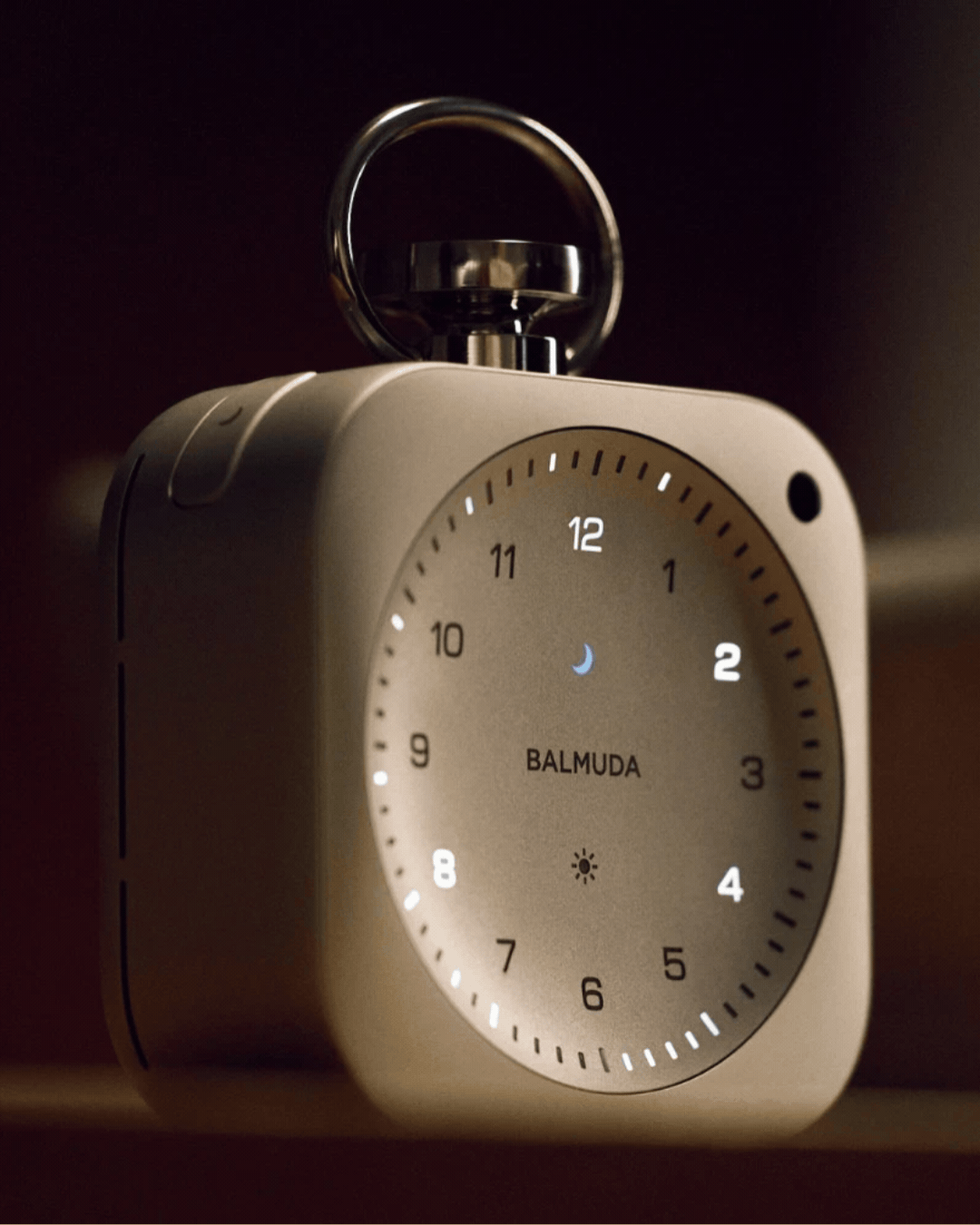 Silver-clad BALMUDA The Clock in a rotating gif, shown at nighttime and against lilac background.