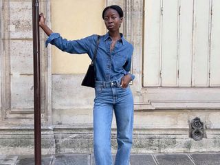 Influencer Sylvie Mus poses on a Paris sidewalk wearing a denim-on-denim look with a jeans button-down shirt, jeans, and a black shoulder bag