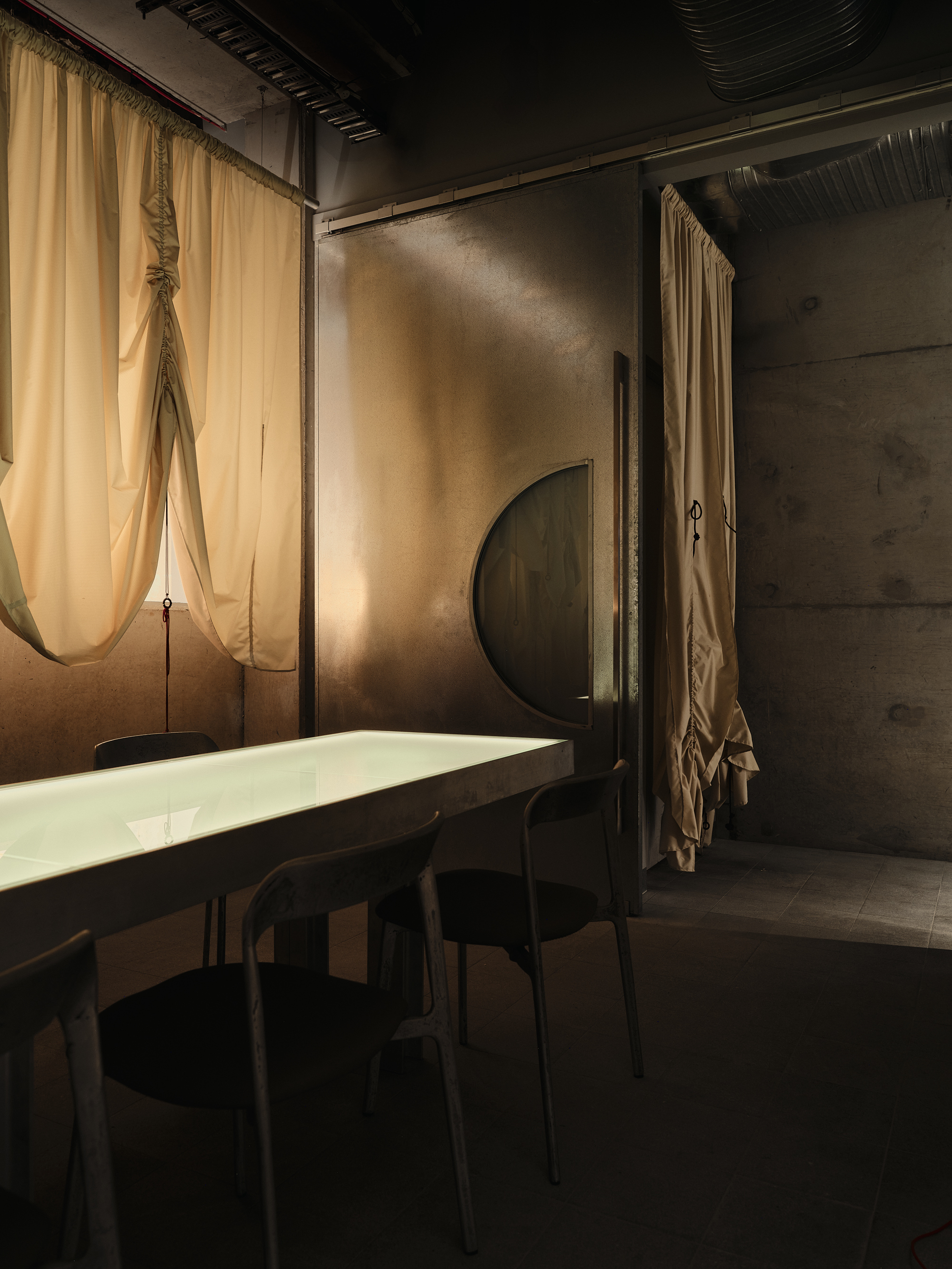 a long dining table with chairs flanking each side in an industrial room with concrete walls and draped curtains