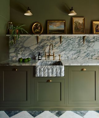 A luxurious kitchen featuring an olive green cabinet base paired with a veined Arabescato marble worktop and backsplash. The standout feature is a marble apron-front sink with brass taps.