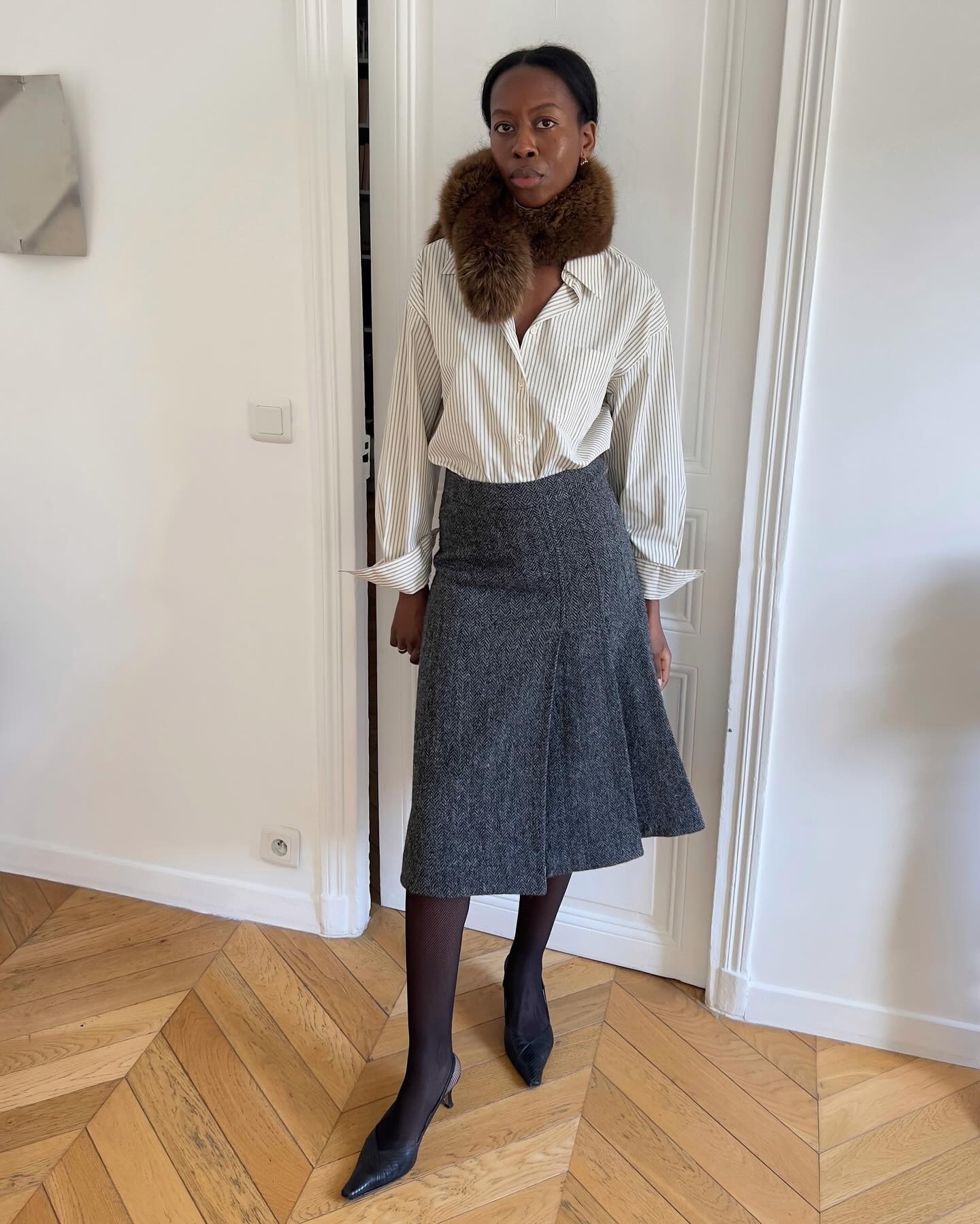 Sylvie wears a fur scarf, striped button-down shirt, gray skirt, sheer tights, and black pumps.