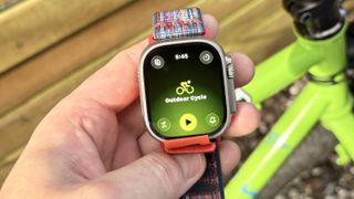 Close-up of the Apple Watch Ultra 3 in a user's hand with the Outdoor Cycle workout tracking mode shown; a lime green bike is out of focus in the distance