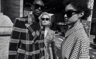 Three models wearing chunky black sunglasses and coats stood near a underpass