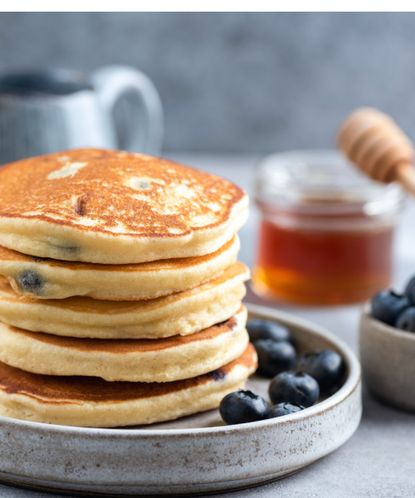 Close up of stack of fluffy pancakes and blueberries in a gray stoneware serving dish, with a pot of honey visible in soft focus in the background 