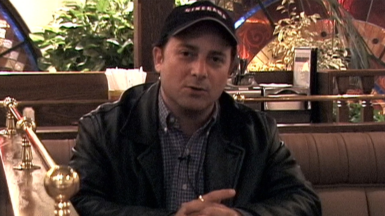 Kevin Pollack wearing a ball cap, sitting in a booth in The Aristocrats.