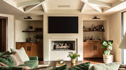 Living room with beige plaster-effect walls and ceiling, light painted ceiling beams, green velvet sofa and open fireplace