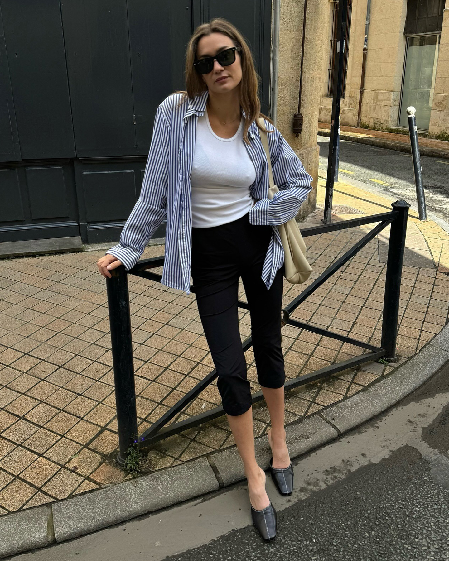 French style influencer Anne-Laure Mais poses on a street corner wearing a striped button-down shirt, white top, black capri pants, and black mule heels