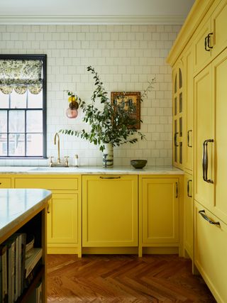 yellow kitchen cabinetry with shaker profile, white tiled backsplash going to the ceiling, white marble counter, timber parquetry floors, and a vase with large green foliage in it on the counter next to the window