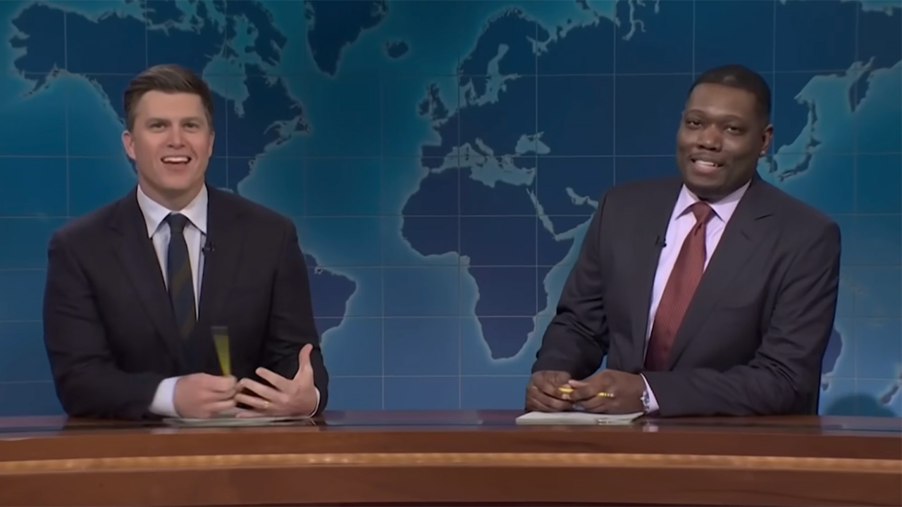 A screenshot of Colin Jost and Michael Che both in suits and ties and sitting at the Weekend Update desk. Che is holding a pencil.