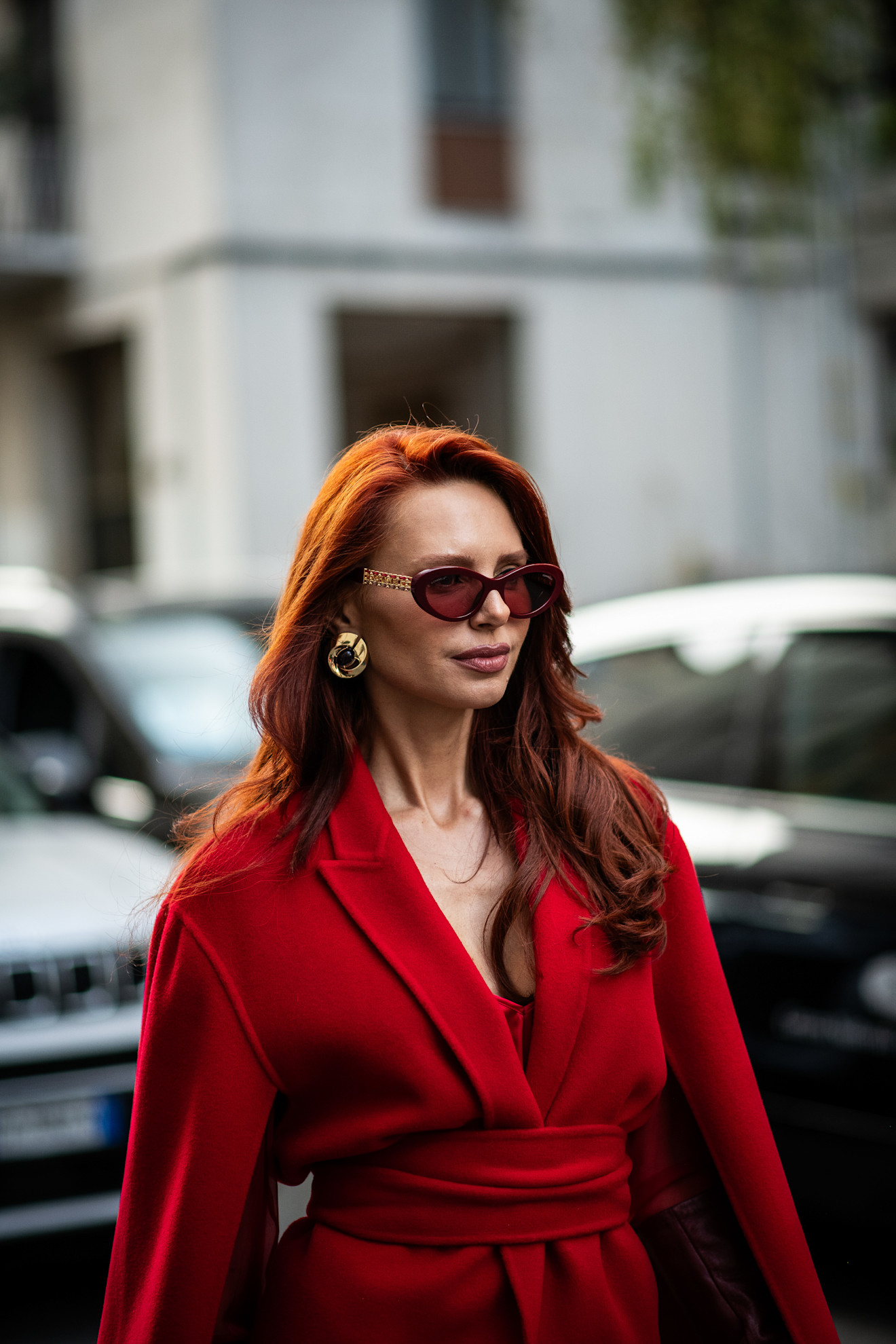 a woman with bright red hair wearing sunglasses, gold earrings and a red wrap coat