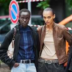 A guest (L) wears a black zipper hood sweater, a blue striped print pattern high neck / turtleneck wool pullover, a white and brown print pattern shoulder bag, a black shiny leather belt, faded blue denim large pants, a guest wears a beige V-neck t-shirt, a brown faded leather jacket, a belt, black denim pants, a guest (R) wears short cropped black hair, a black leather shoulder bag with a chain strap and gold key charm, a long black wool coat with notched lapels, a white T-shirt with a navy crew neckline, indigo straight-leg denim jeans fastened with a brown leather belt with a large gold rectangular buckle, outside Sportmax, during Milan Fashion Week - Womenswear Spring/Summer 2026, on September 26, 2025 in Milan, Italy