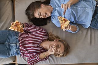 getting over a breakup - Two laughing young women lying down eating pizza together
