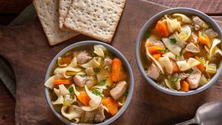 Two bowls of chicken noodle soup with vegetables and dry crackers