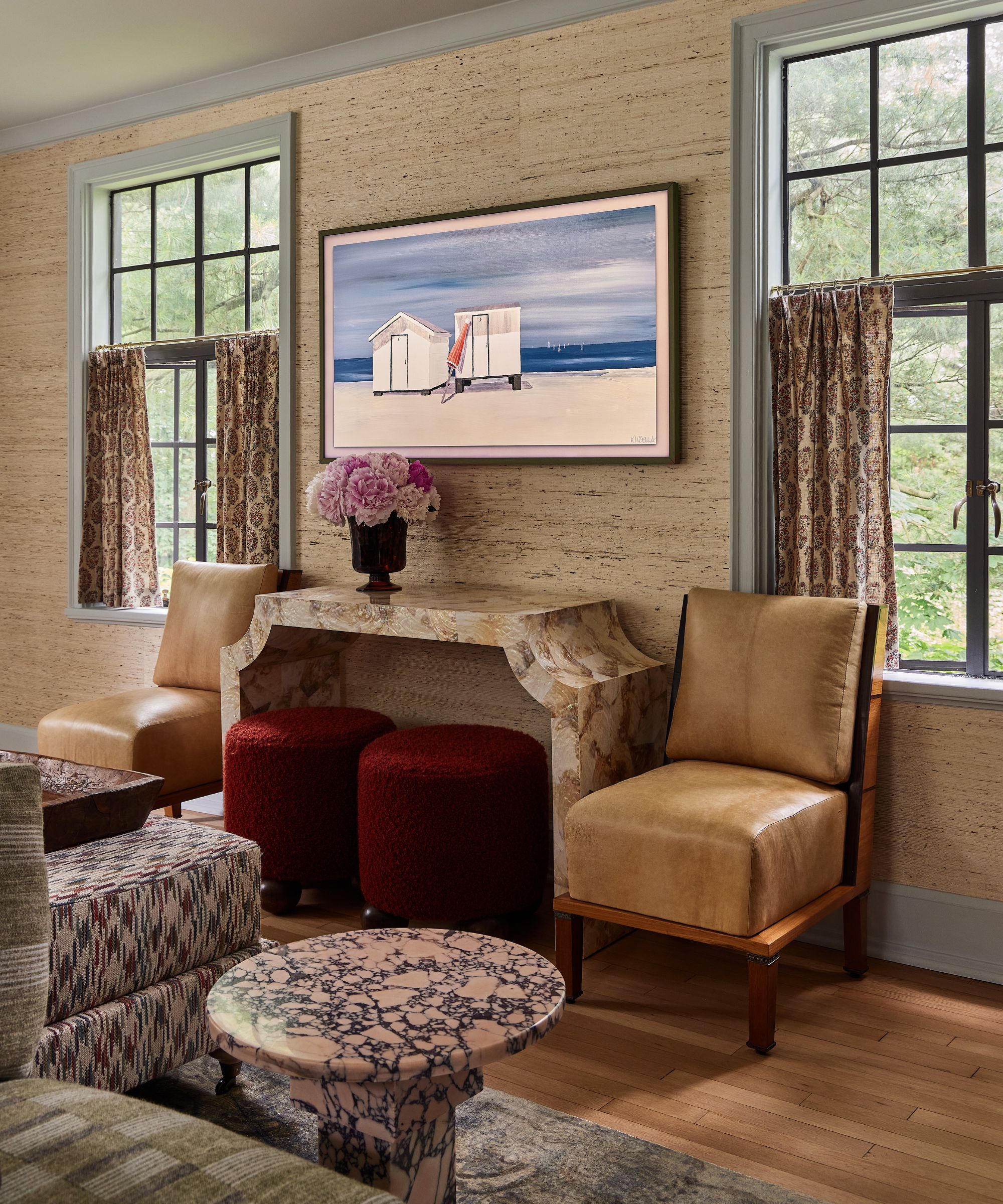 a living room with grasscloth walls, two blue trimmed windows, with a marble console table vignette with vintage chairs and red ottomans