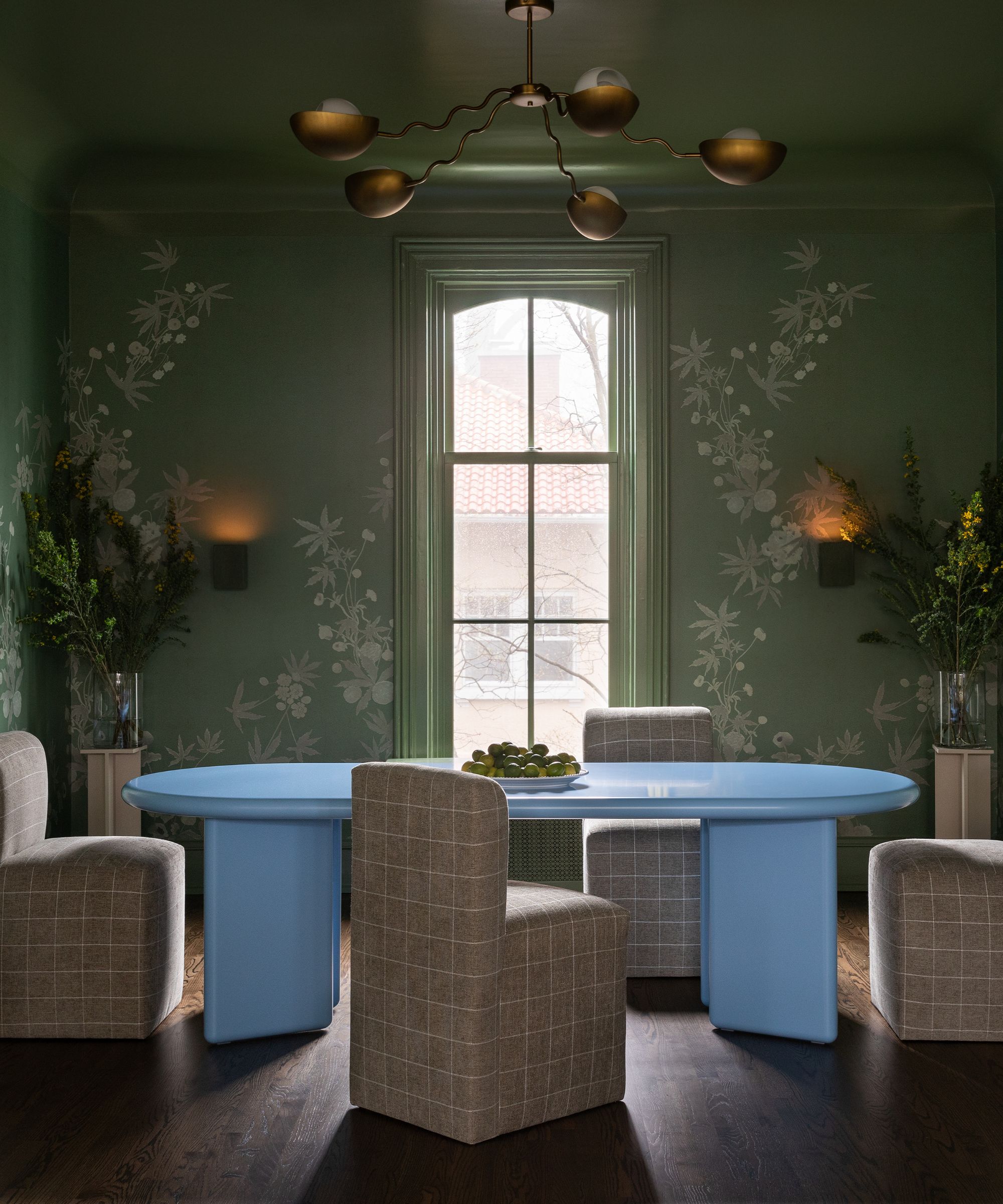 A moody dining room featuring sage green walls with white floral botanical patterns. A large, matte light-blue oval table sits in the center, surrounded by grey checkered upholstered chairs. A modern brass multi-arm chandelier hangs from the green ceiling, and a tall window provides soft natural light.