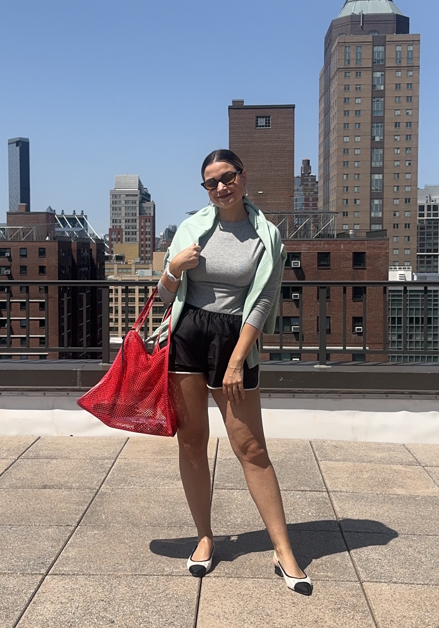 Writer Irina Grechko wearing black track shorts with gray long-sleeve top, mint sweater thrown over the shoulder, red mesh tote bag, and black-toe white slingback flats.