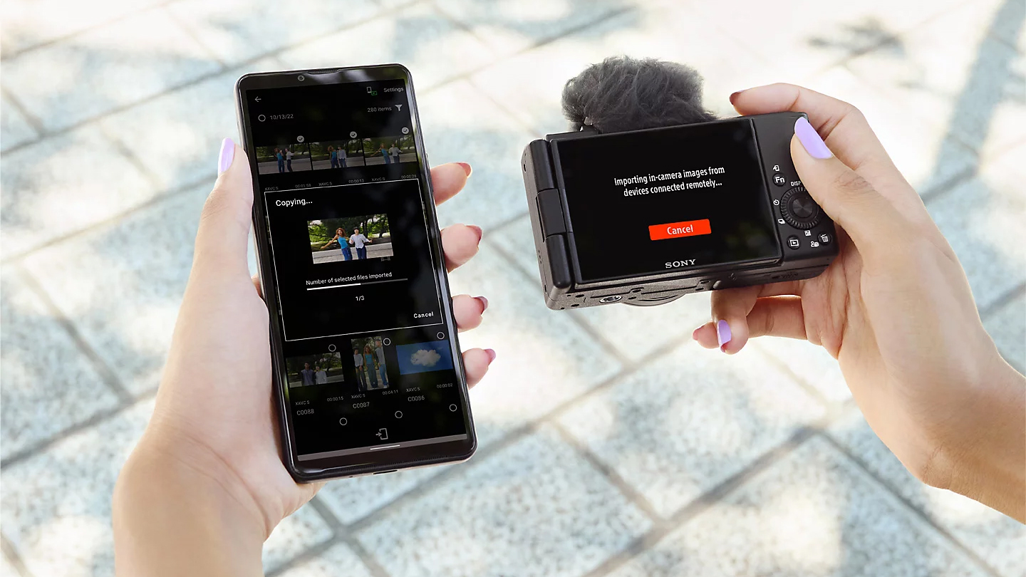 A person holds a smartphone displaying a file transfer screen while using a Sony ZV-1F camera to import images