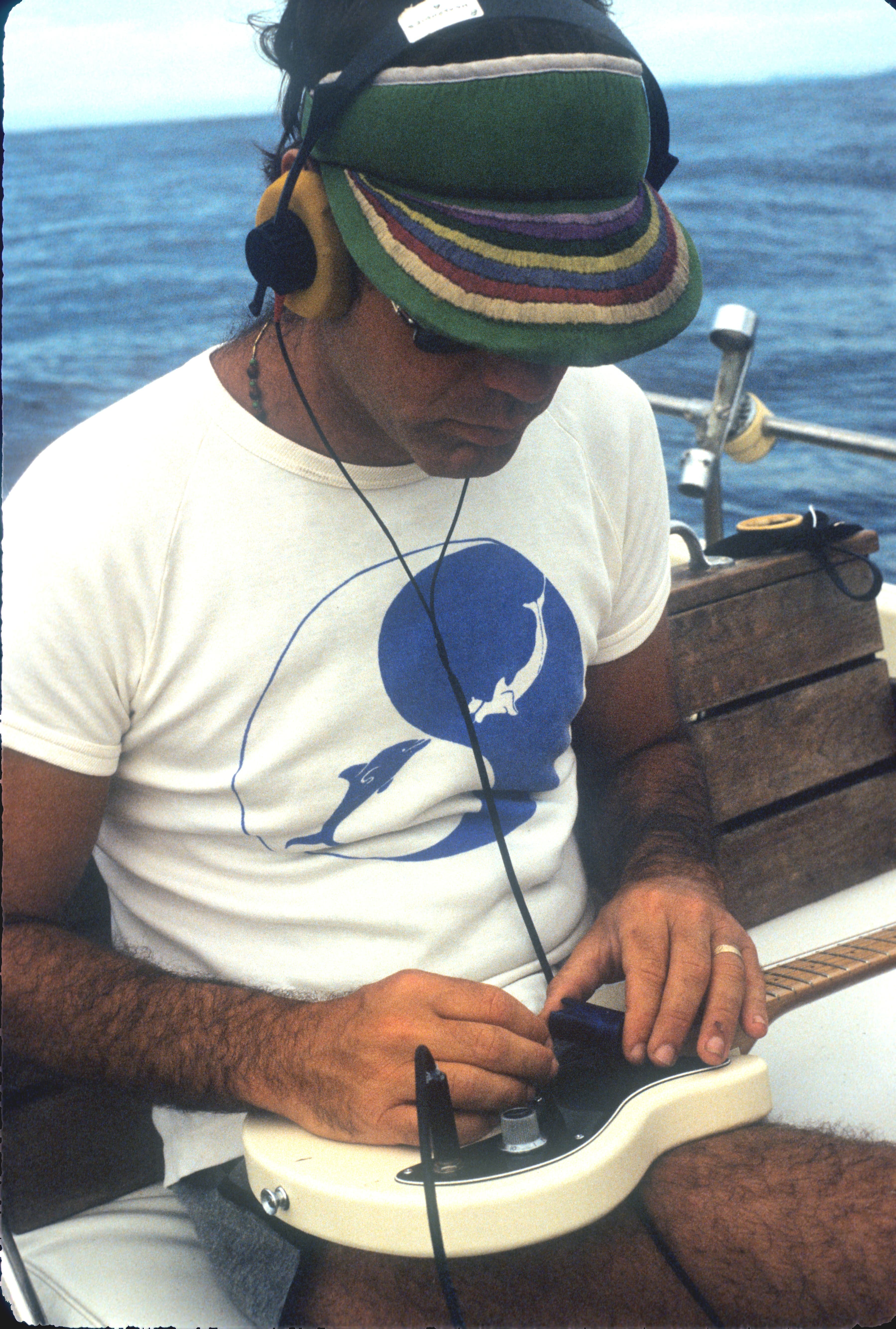 A man playing a guitar with headphones in, on a boat in on the ocean.