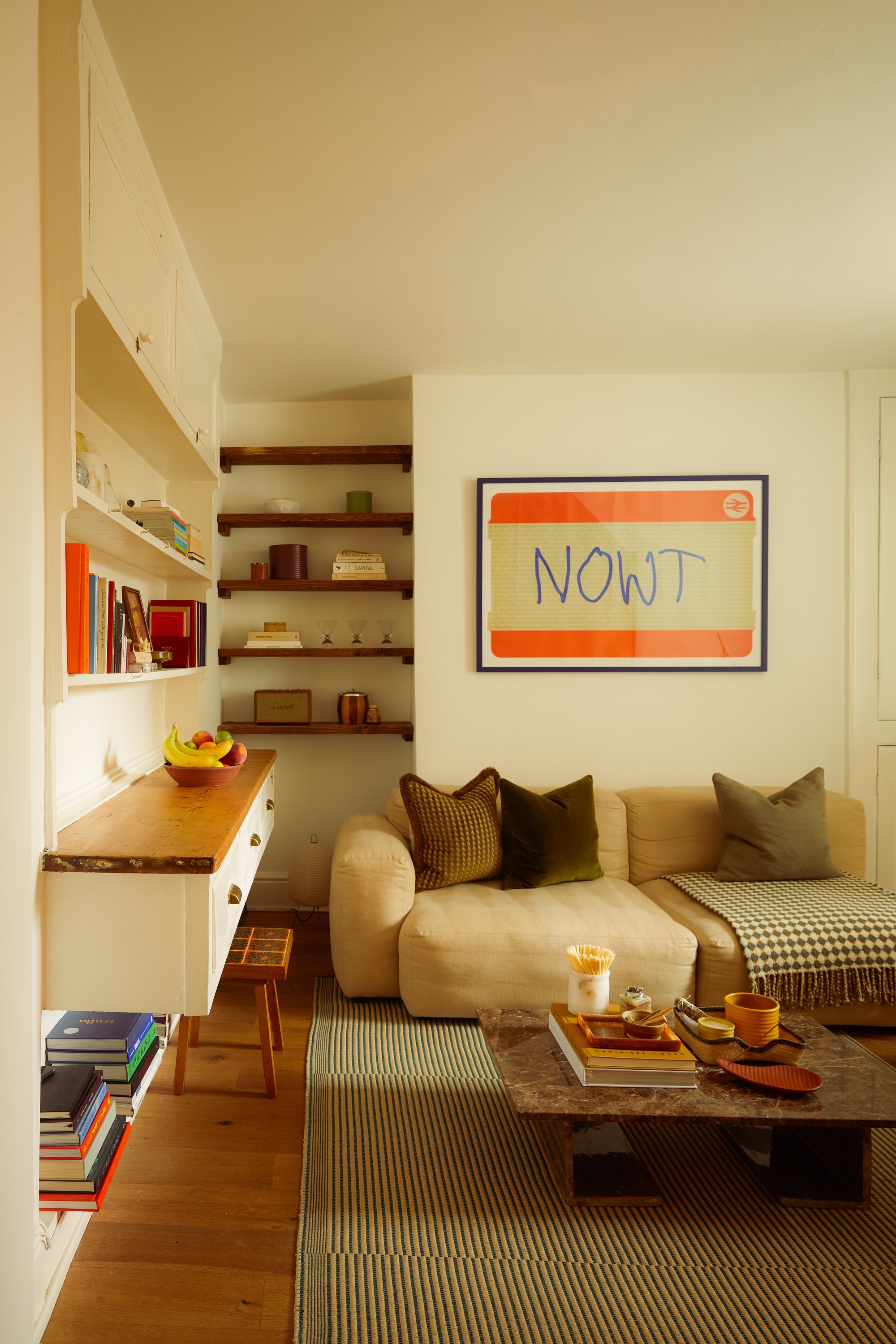 Image of a warm, cream living room with a cozy beige sofa that has green throw pillows. There is a green area rug, a brown marble coffee table, and wooden shelves on the walls.