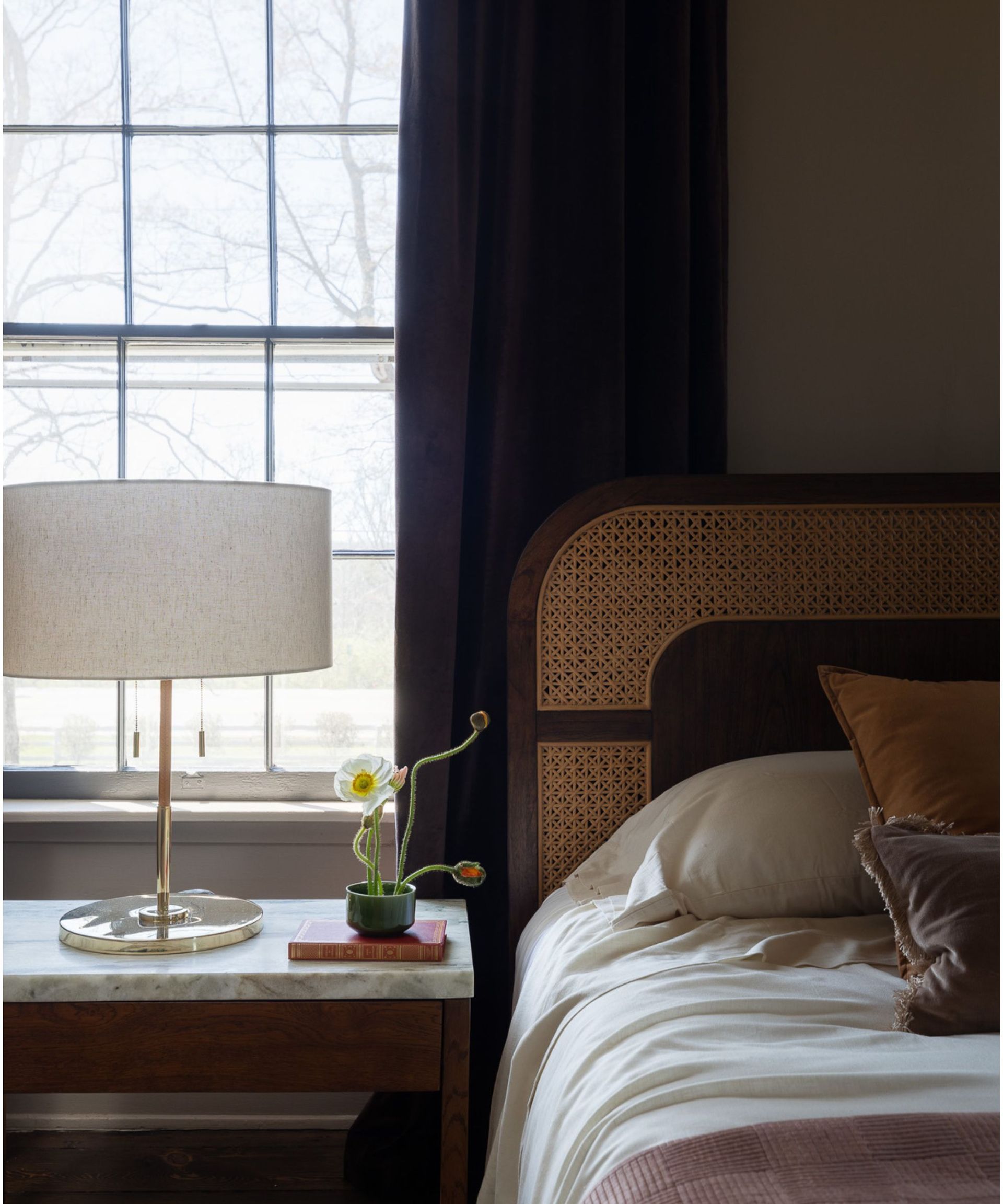 close up of a bed in a small bedroom painted in a soft brown color with a marble topped bedside styled with a lamp and small floral display