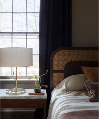 close up of a bed in a small bedroom painted in a soft brown color with a marble topped bedside styled with a lamp and small floral display