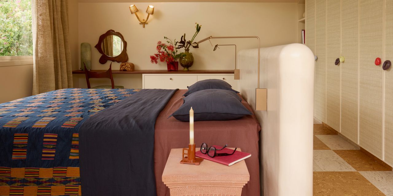 A beige bedroom with a checkerboard terrazzo floor, and a bed with a white headboard that stands out from the wall. The bedding is a combination of wine and plum colors with a blue pattered quilt on top.