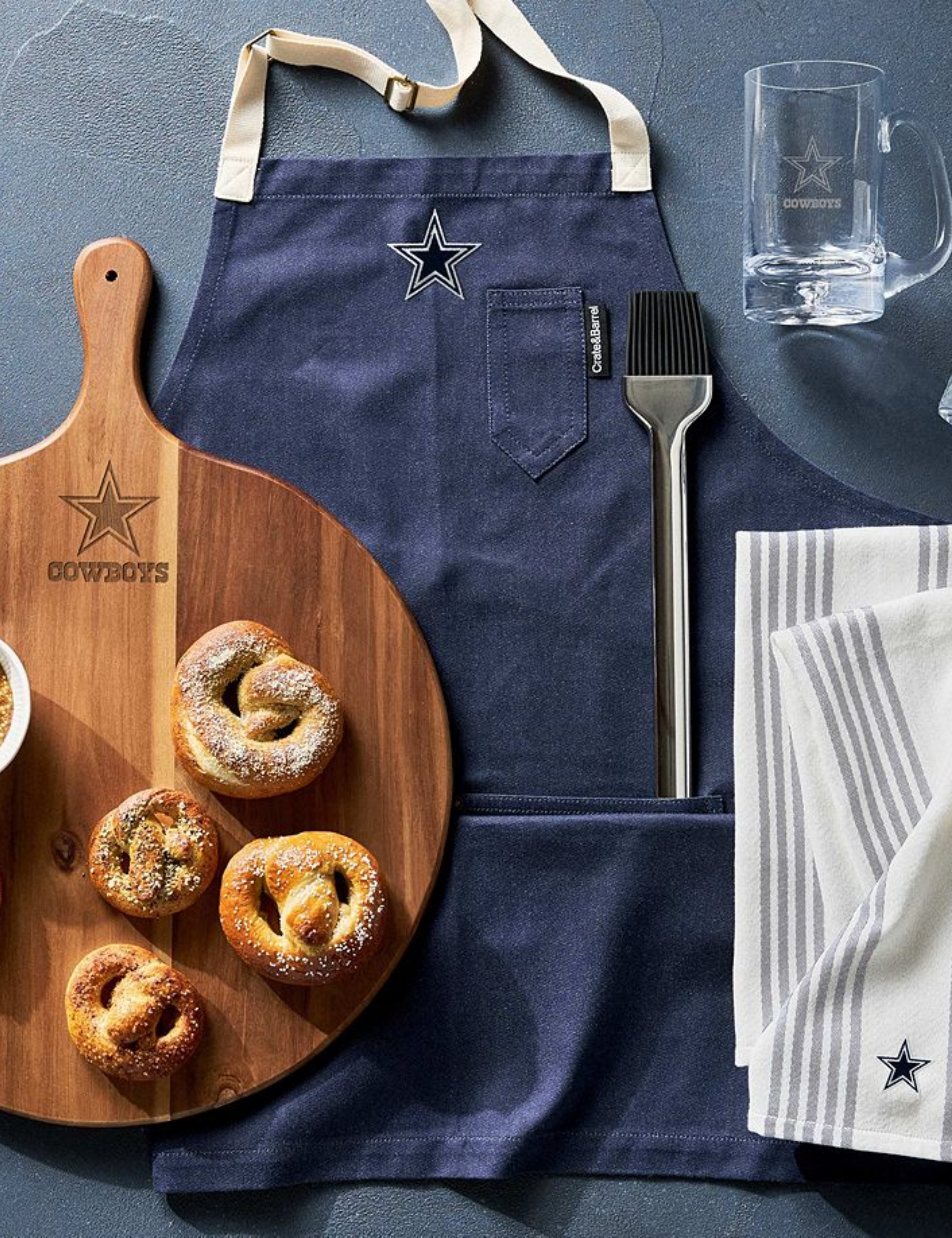 Crate &amp;amp; Barrel x NFL-branded wooden serving board with soft pretzels, apron, BBQ tool, beer mug, and dish towels pictured from a bird's eye view.