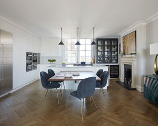 A large white kitchen with herringbone flooring and dark blue chairs, broken up by L-shaped units.