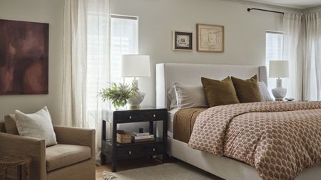 Bedroom with bed, two bedside tables with lamps and books, and armchair in beige/red color scheme. Two windows with open curtains.