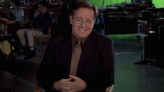 Phil Hartman smiles while talking in a director's chair in NewsRadio.
