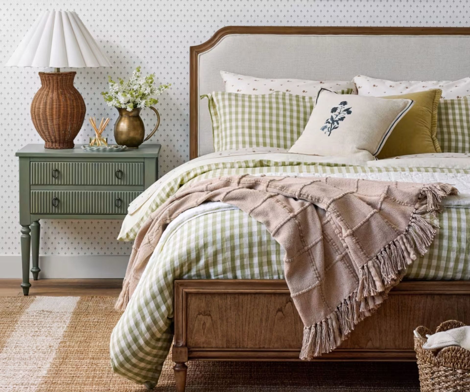 magnola styled bedroom with floral white and blue wallpaper, a wooden bedframe with a linen headboard, green gingham bed linens and a green nightstand with rattan scalloped lamp