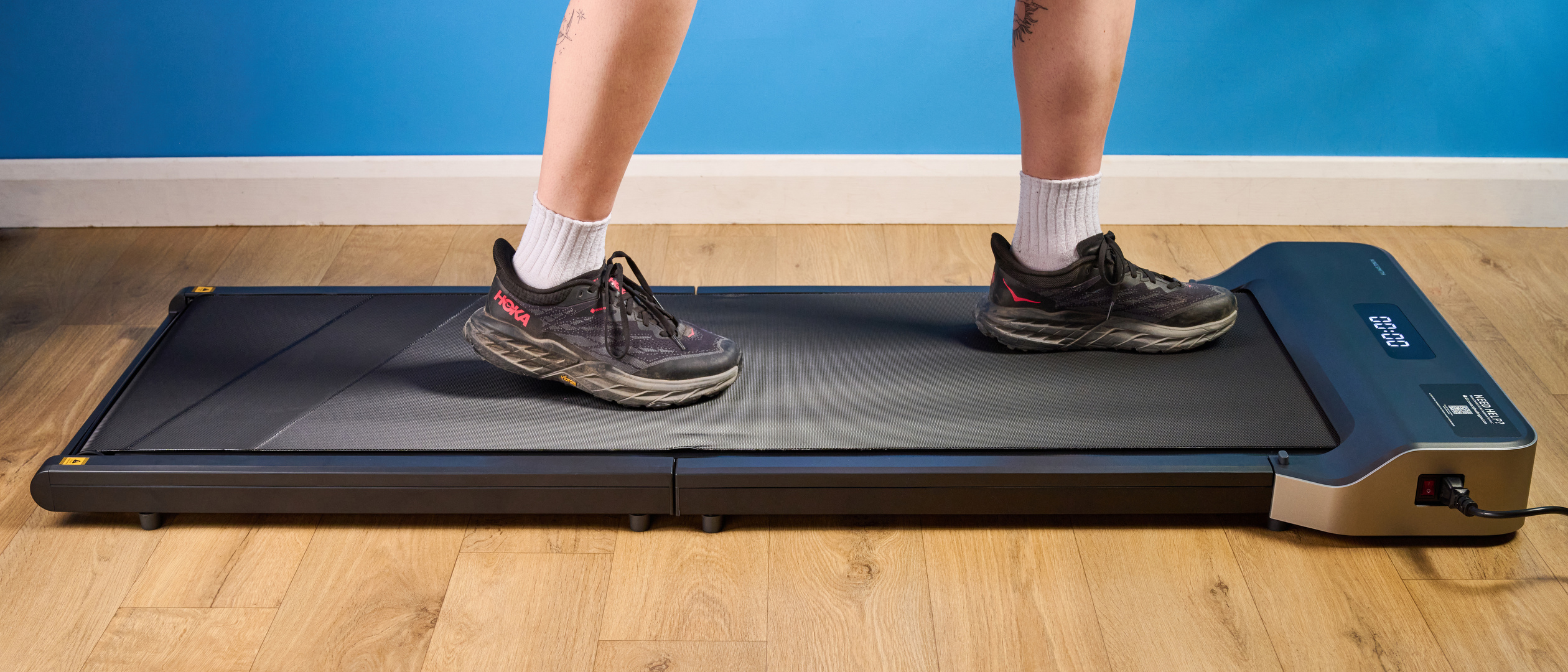 A woman walking on the WalkingPad Z1 wearing Hoka running shoes photographed in front of a blue background. 