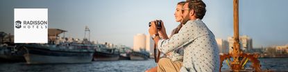 Radission Hotels logo placed on a wide image of a couple taking pics from a river side