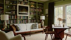 a green living room and home office with a bookshelf and dark green curtains. a couch is flanked by matching table lamps