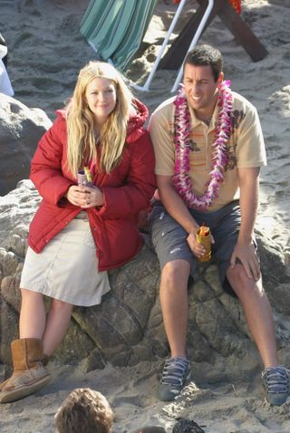 Drew Barrymore has long blonde hair and is wearing a big red coat as she films 50 First Dates on the beach with Adam Sandler