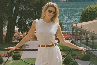 Woman wearing white muscle tee, white pants, brown belt, and sunglasses in Lake Como, Italy.