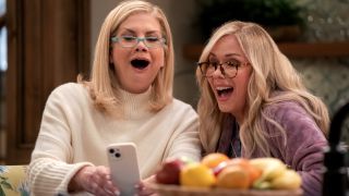 (L to R) Kristen Johnston as Carol and Leanne Morgan as Leanne smiling while looking at a phone.