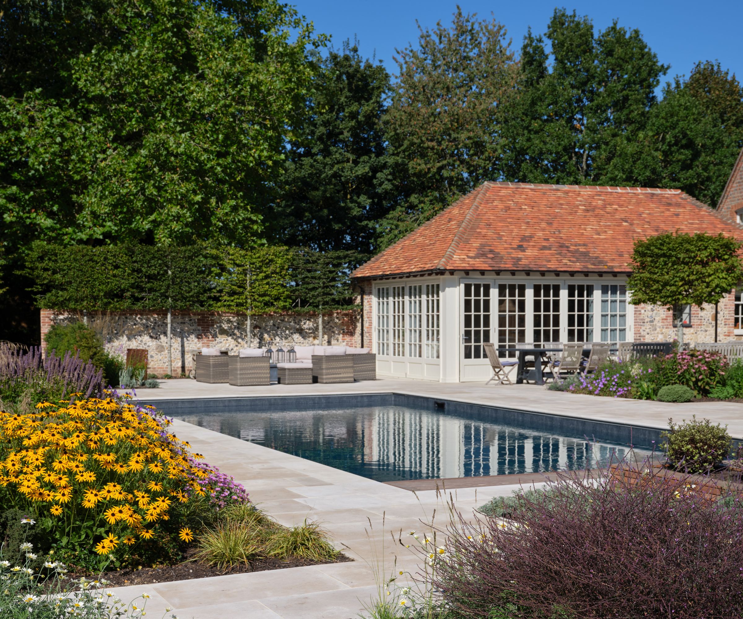 A country garden swimming pool with limestone surround, wicker garden furniture, rudbeckia and lavender borders, and a white-painted pool house with terracotta roof.