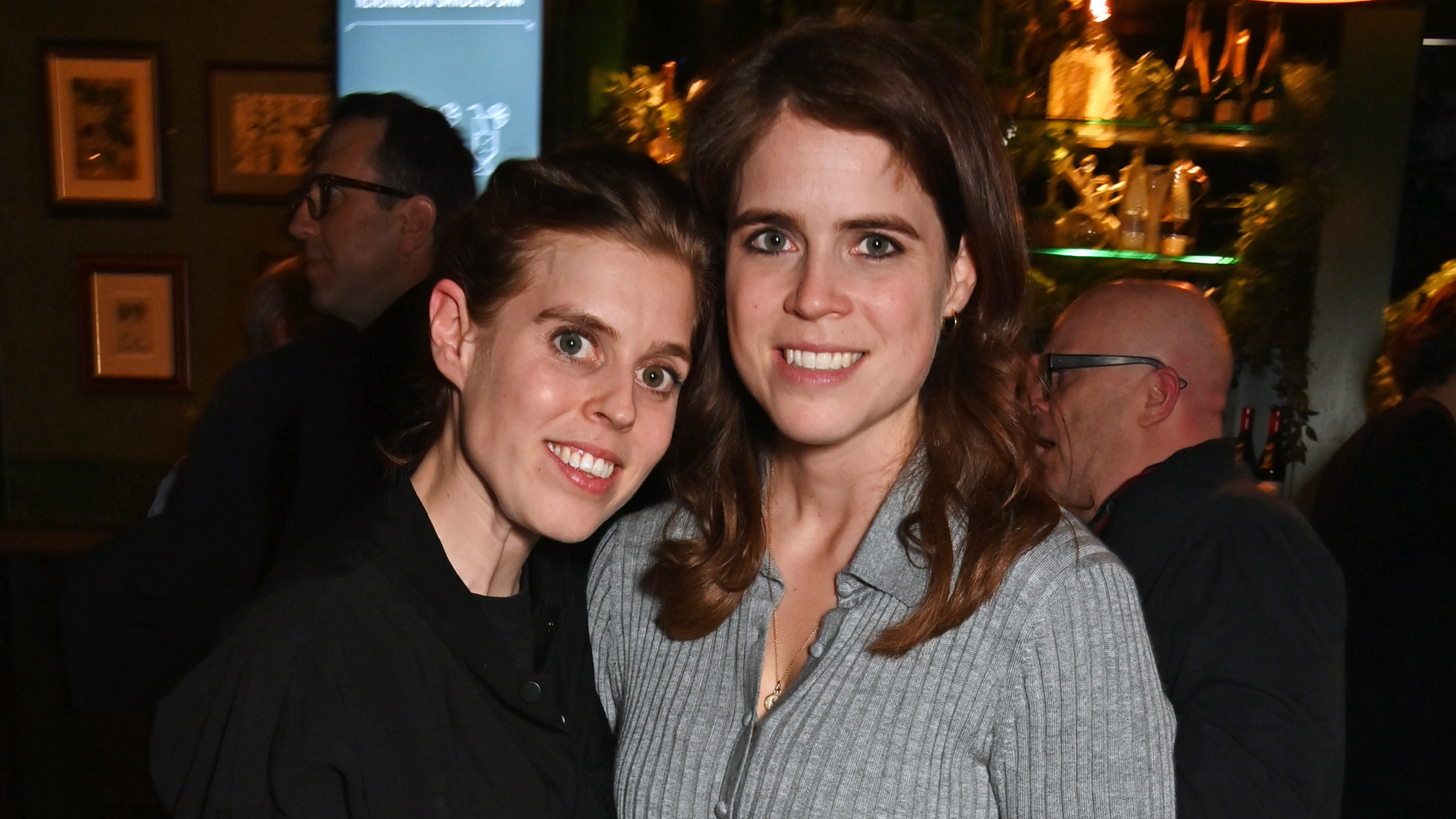 Princess Beatrice wearing black and Princess Eugenie in a gray dress posing in a restaurant