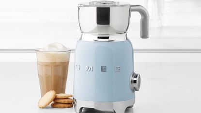 smeg milk frother in pastel blue on a countertop beside a latte and some biscuits