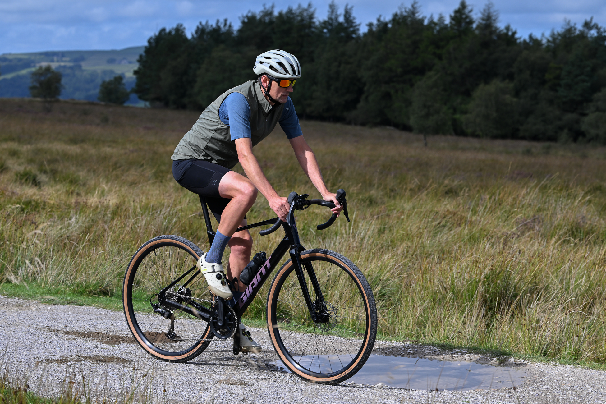 man riding a black gravel bike wearing black shorts, blue socks, blue jersey and green gilet with beige SPD shoes on a gravel trail