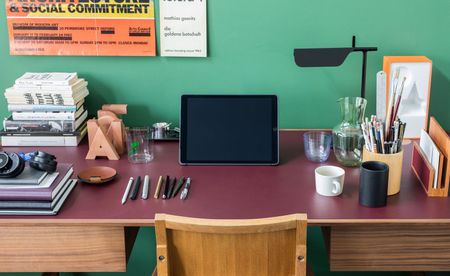 Wooden chair and wine coloured wooden desk with a tablet, a lamp, pens, books, a white mug and glassware against a green wall