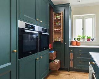 Kitchen with dark green cabinetry, slim pantry slide open, sink and kitchen window, wooden flooring.