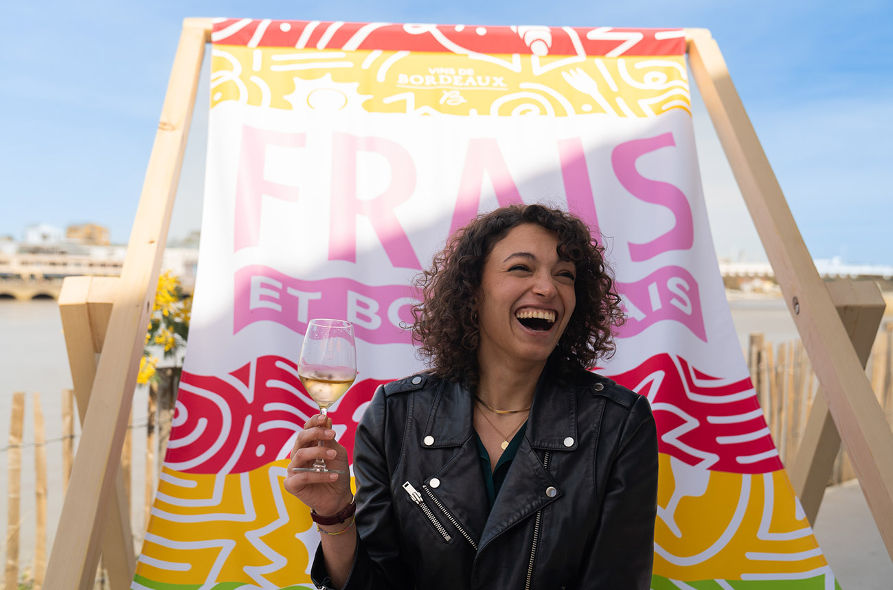 A lady smiling holding a glass of white wine, with 'Frais et Bordelais' on a flag in the background