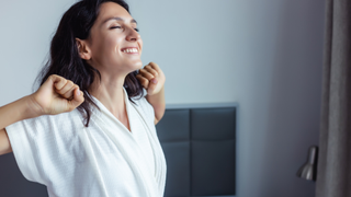 A woman wearing a white robe stretches while smiling in the morning.