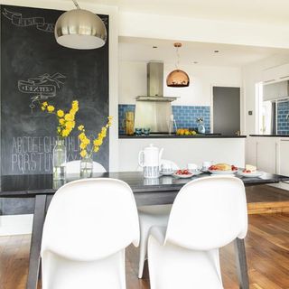 open plan kitchen and dining area with wooden floorboards, a blue brick splashback in the kitchen, wall with blackboard paint and a black dining table with white chairs